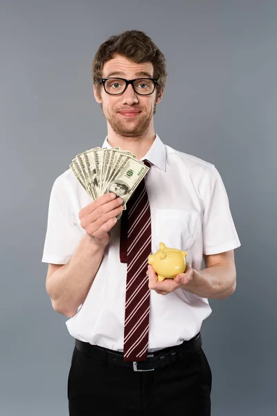 Businessman in glasses holding piggy bank and dollar banknotes on grey background — Stock Photo