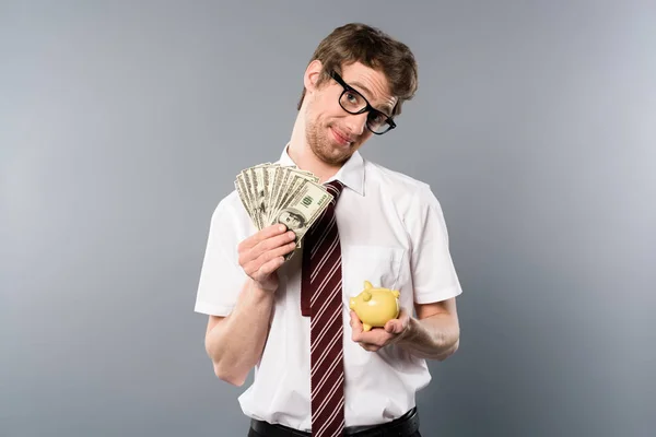 Confused businessman in glasses holding piggy bank and dollar banknotes on grey background — Stock Photo
