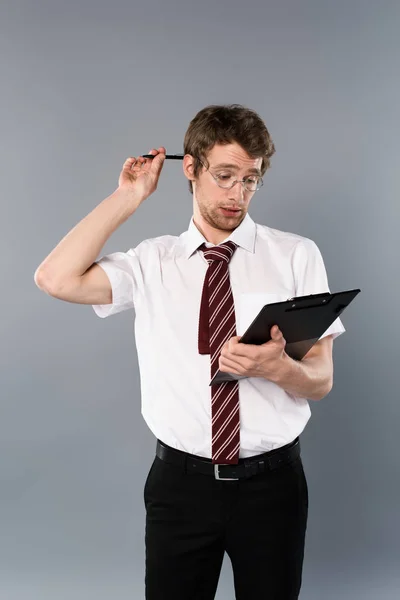 Thoughtful man with pen holding clipboard on grey background — Stock Photo