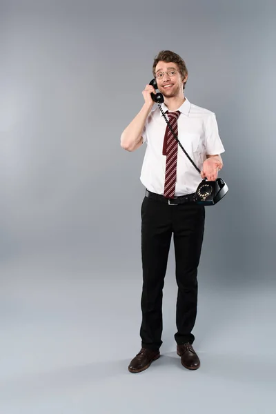 Улыбающийся бизнесмен в очках разговаривает по винтажному телефону на сером — стоковое фото
