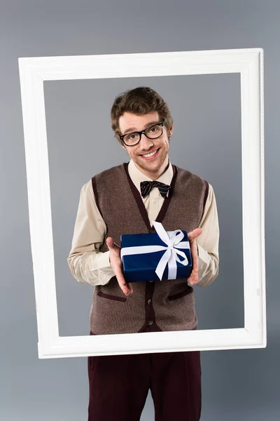 Hombre sonriente en marco blanco sosteniendo caja de regalo sobre fondo gris - foto de stock