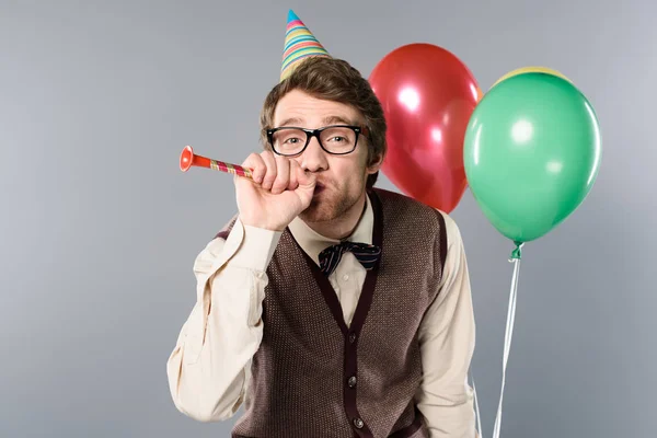 Funny man in glasses and party cap holding multicolored balloons and blowing in party horn on grey background — Stock Photo