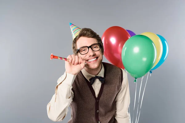 Smiling man in glasses and party cap holding multicolored balloons and party horn on grey background — Stock Photo