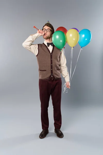 Mann mit Brille und Partymütze hält bunte Luftballons in der Hand und bläst in Partyhorn auf grauem Hintergrund — Stockfoto