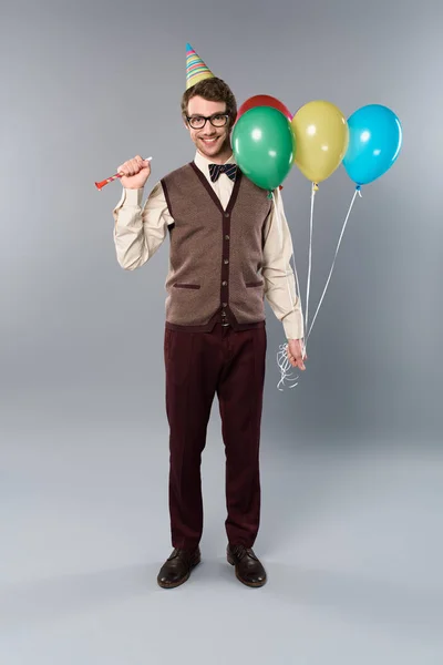 Glücklicher Mann mit Brille und Partymütze mit bunten Luftballons und Partyhorn auf grauem Hintergrund — Stockfoto