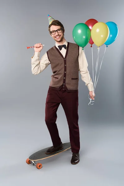 Glücklicher Mann mit Brille und Partymütze mit bunten Luftballons und Partyhorn auf Longboard vor grauem Hintergrund — Stockfoto