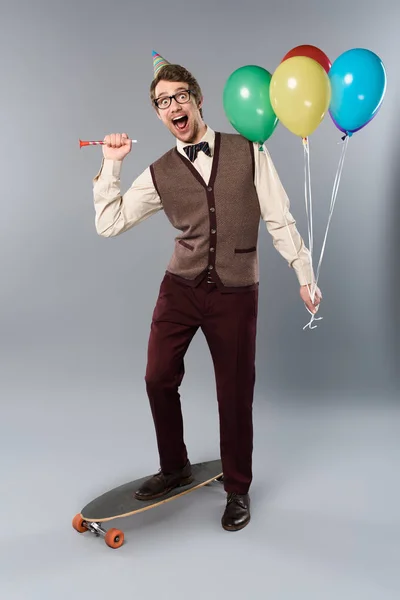 Excited man in glasses and party cap holding multicolored balloons and party horn while standing on longboard on grey background — Stock Photo