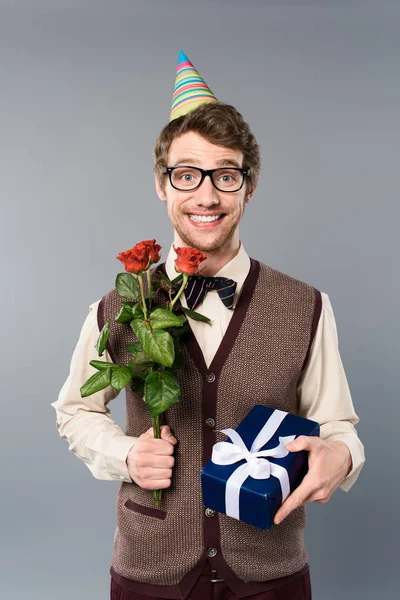 Усміхнений чоловік у вечірній кепці тримає букет з троянд та подарункової коробки ізольовано на сірому — стокове фото