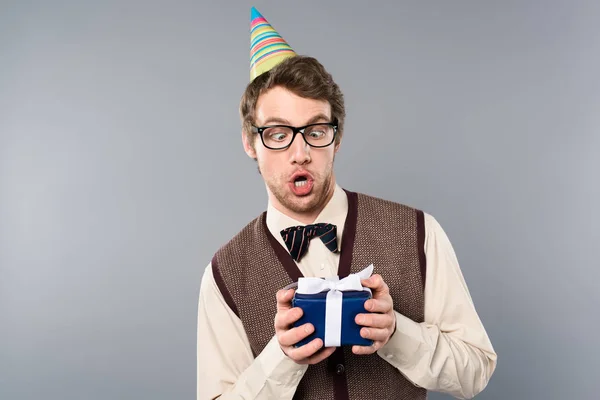 Schockierter Mann mit Party-Mütze hält Geschenkbox auf Grau — Stockfoto