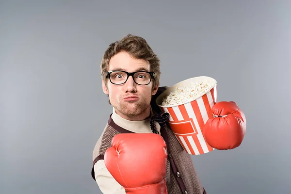 Angry man in boxing gloves holding bucket of popcorn on grey background — Stock Photo