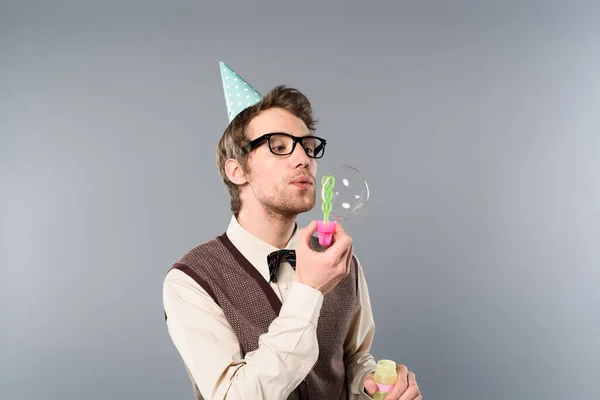 Homme en vêtements vintage et bonnet de fête soufflant des bulles de savon sur fond gris — Photo de stock