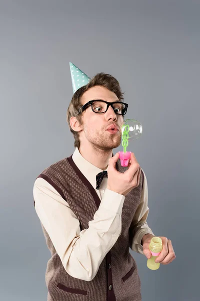 Homme drôle en vêtements vintage et chapeau de fête soufflant des bulles de savon sur fond gris — Photo de stock