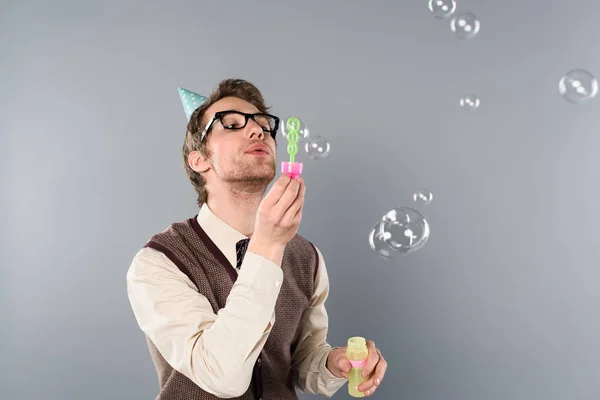 Homme adulte en vêtements vintage et bonnet de fête soufflant des bulles de savon sur fond gris — Photo de stock