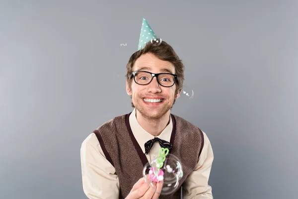 Улыбающийся человек в винтажной одежде и кепке для вечеринок выдувает мыльные пузыри на сером фоне — стоковое фото