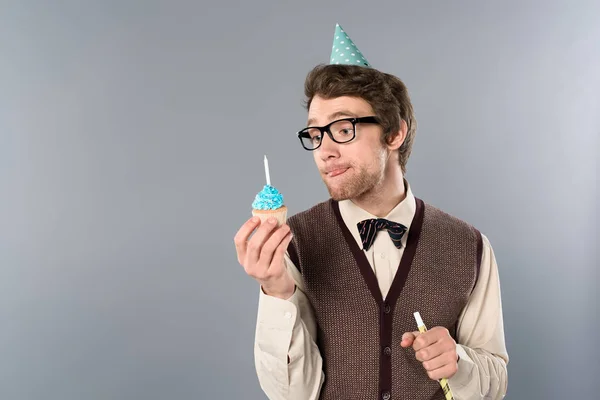 Homme dans des lunettes et casquette de fête avec expression drôle visage tenant cupcake avec bougie — Photo de stock