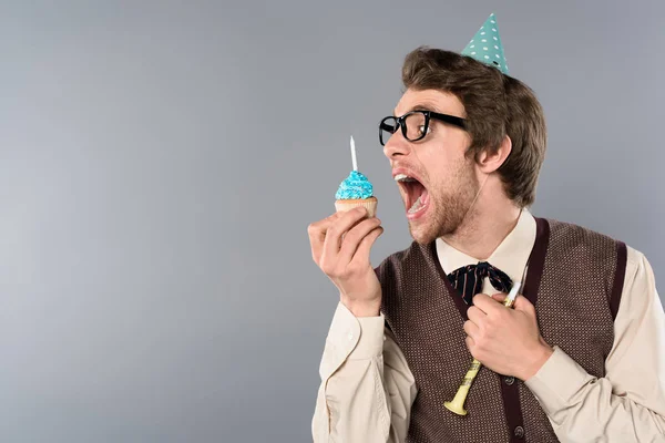 Man in glasses and party cap with funny face expression eating cupcake with candle — Stock Photo
