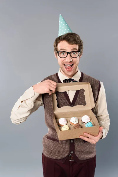 Homem animado em óculos e tampa de festa segurando caixa com cupcakes — Fotografia de Stock