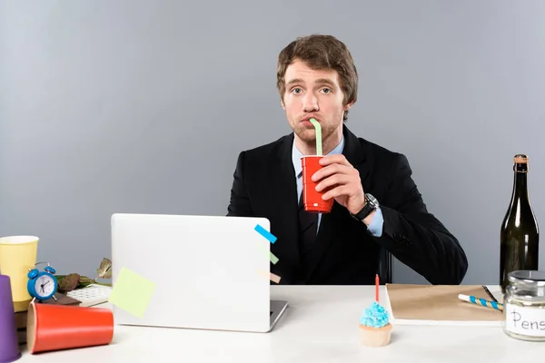 Geschäftsmann sitzt mit Cupcake am Arbeitsplatz und trinkt aus Pappbecher isoliert auf grau — Stockfoto