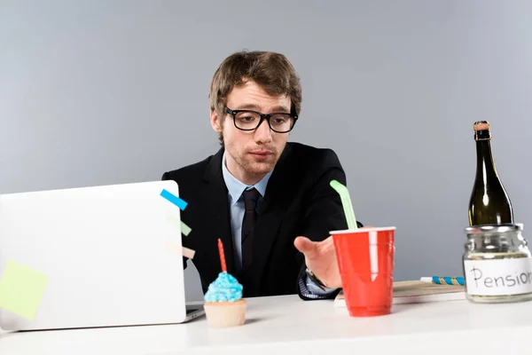 Tired businessman sitting at workplace with cupcake and looking at paper cup isolated on grey — Stock Photo