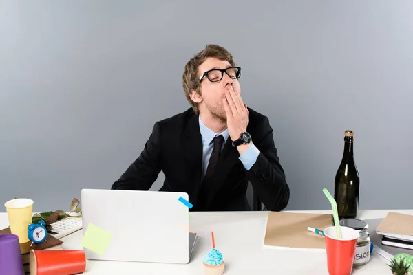 Бизнесмен зевает на рабочем месте возле кексов и бумажных стаканчиков, изолированных на сером — стоковое фото