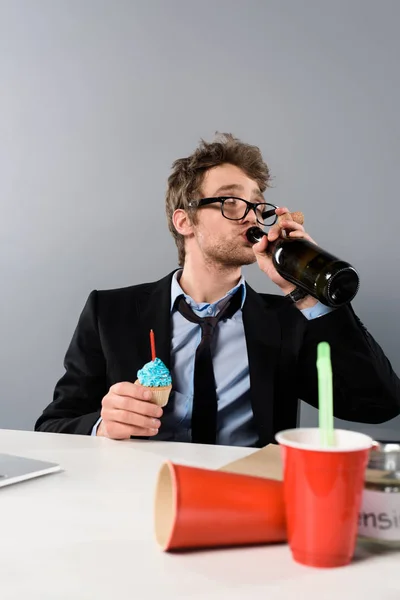 Пьяный бизнесмен, сидящий на рабочем месте и пьющий шампанское из бутылки, изолированной на сером — стоковое фото