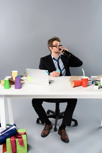Betrunkener Geschäftsmann sitzt am Arbeitsplatz und trinkt Champagner aus Flasche in der Nähe von Geschenkboxen — Stockfoto