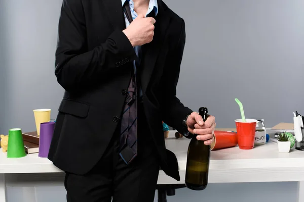 Cropped view of drunk businessman standing near workplace with bottle of champagne — Stock Photo