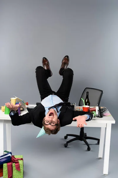 Happy businessman in party cap lying on table at messy workplace — Stock Photo