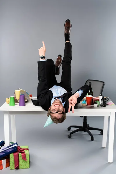 Glücklicher Geschäftsmann in Partymütze, der am unordentlichen Arbeitsplatz auf dem Tisch liegt und Friedenszeichen zeigt — Stockfoto