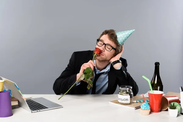 Drunk businessman in birthday cap smelling rose at workplace — Stock Photo