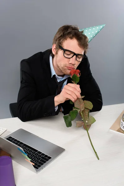 Drunk businessman in birthday cap smelling rose at workplace on grey background — Stock Photo