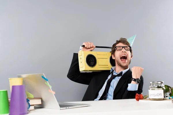 Happy businessman sitting at workplace in party cap with vintage tape recorder and rejoicing on grey background — Stock Photo