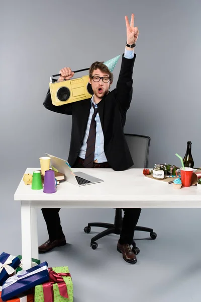 Homme d'affaires ivre assis sur le lieu de travail en bonnet de fête avec magnétophone vintage sur fond gris — Photo de stock