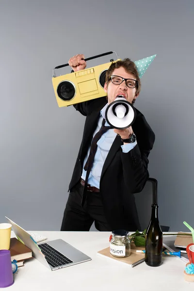 Empresario borracho en el lugar de trabajo con gorra de fiesta con grabadora vintage gritando en megáfono sobre fondo gris - foto de stock