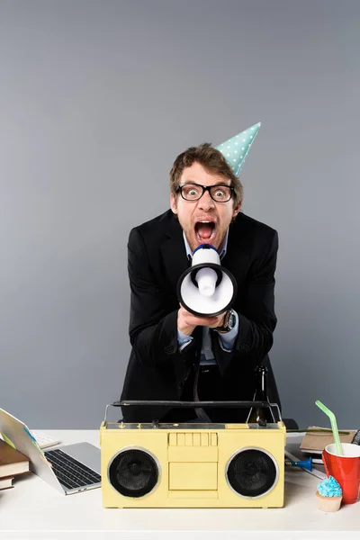 Angry businessman at workplace in party cap near vintage tape recorder screaming in megaphone on grey background — Stock Photo