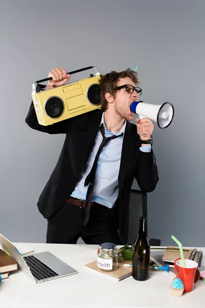 Angry businessman at workplace in party cap holding vintage tape recorder and screaming in megaphone on grey background — Stock Photo