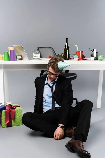 Drunk businessman in party cap sleeping at messy workplace near presents — Stock Photo
