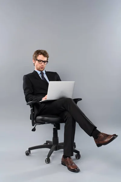 Бизнесмен в костюме сидит в кресле и использует ноутбук на сером фоне — стоковое фото