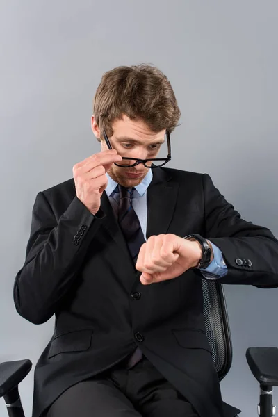 Hombre de negocios en traje sentado en silla y mirando reloj aislado en gris - foto de stock