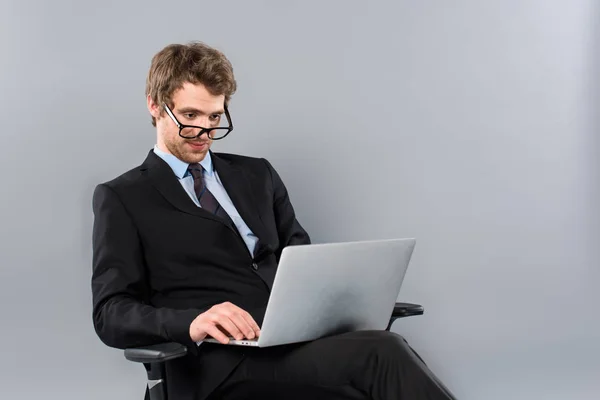 Businessman in suit and glasses sitting in chair and using laptop on grey background — Stock Photo