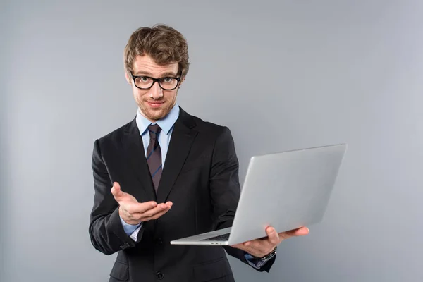 Uomo d'affari in giacca e cravatta con computer portatile e gesti su sfondo grigio — Foto stock