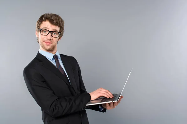 Gracioso hombre de negocios en traje usando portátil sobre fondo gris - foto de stock