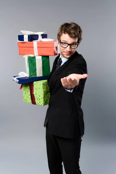 Усміхнений бізнесмен тримає подарунки і жестикулює на сірому фоні — стокове фото
