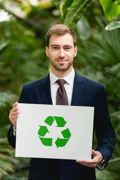 Apuesto hombre de negocios sonriente en traje con tarjeta con signo de reciclaje verde en invernadero - foto de stock