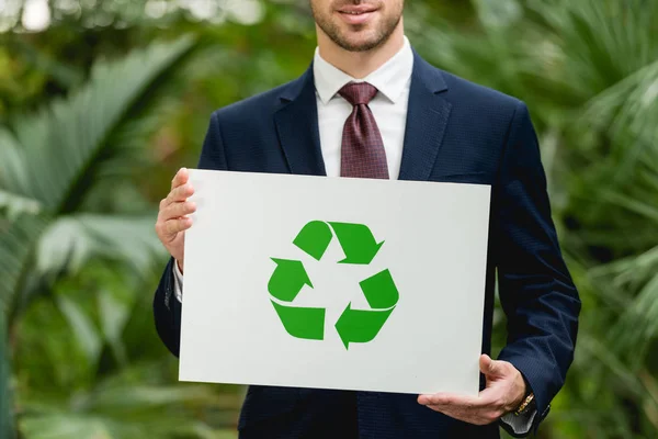 Cropped view of smiling businessman in suit holding card with green recycling sign in greenhouse — Stock Photo