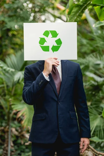 Businessman in suit holding card with green recycling sign in front of face in greenhouse — Stock Photo
