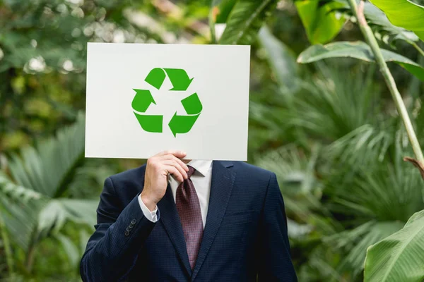 Businessman holding white card with green recycling sign in front of face in greenhouse — Stock Photo