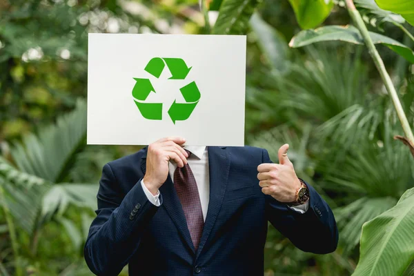 Hombre de negocios con tarjeta blanca con letrero de reciclaje verde en frente de la cara y mostrando el pulgar hacia arriba en el invernadero - foto de stock