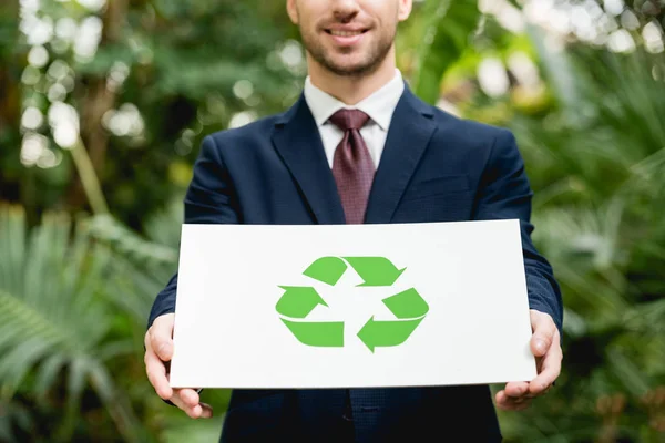 Partial view of smiling businessman in suit holding card with green recycling sign in greenhouse — Stock Photo