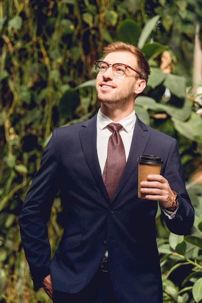 Uomo d'affari sorridente in giacca, cravatta e bicchieri che tiene il caffè per andare in serra — Foto stock